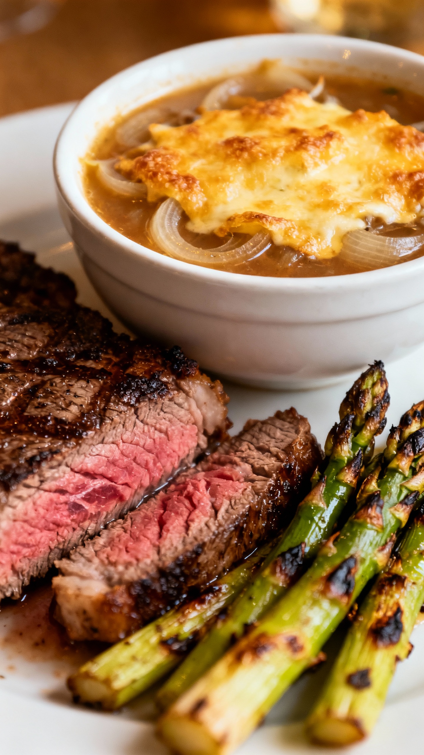 Closeup of onion soup with melted cheese crust, ribeye medium-rare, blistered asparagus