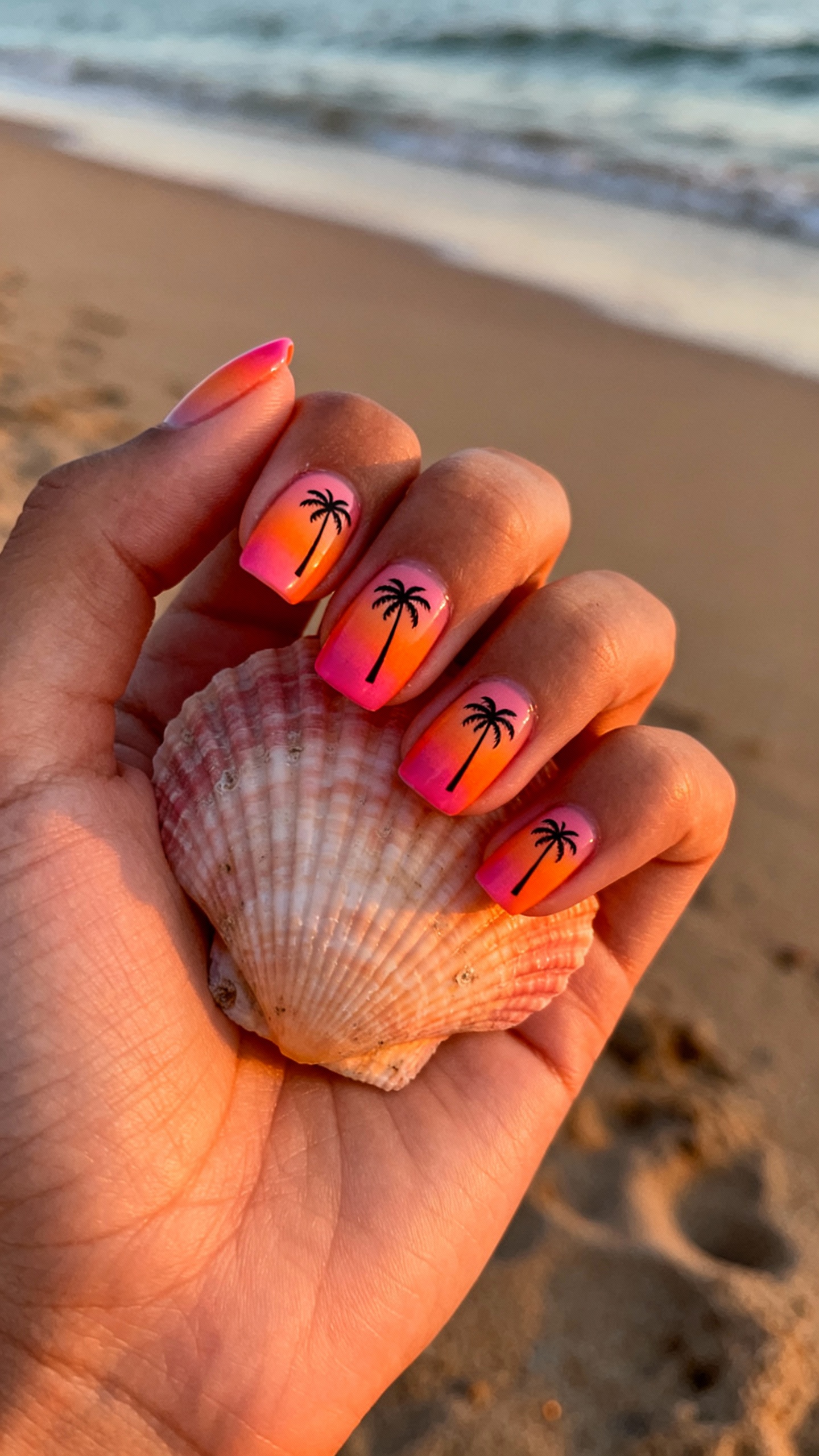 Closeup sunset ombre nails with tiny palm decal, hand holding seashell, sandy shoreline